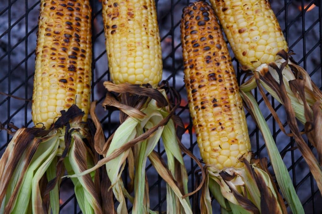 four ears of corn with husks attached charring on a grill over hot coals