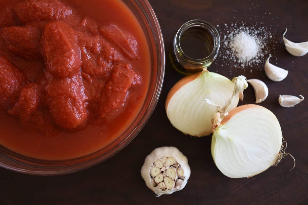 bowl of whole peeled tomatoes on a cutting board with a head of garlic, yellow onion, salt, and olive oil