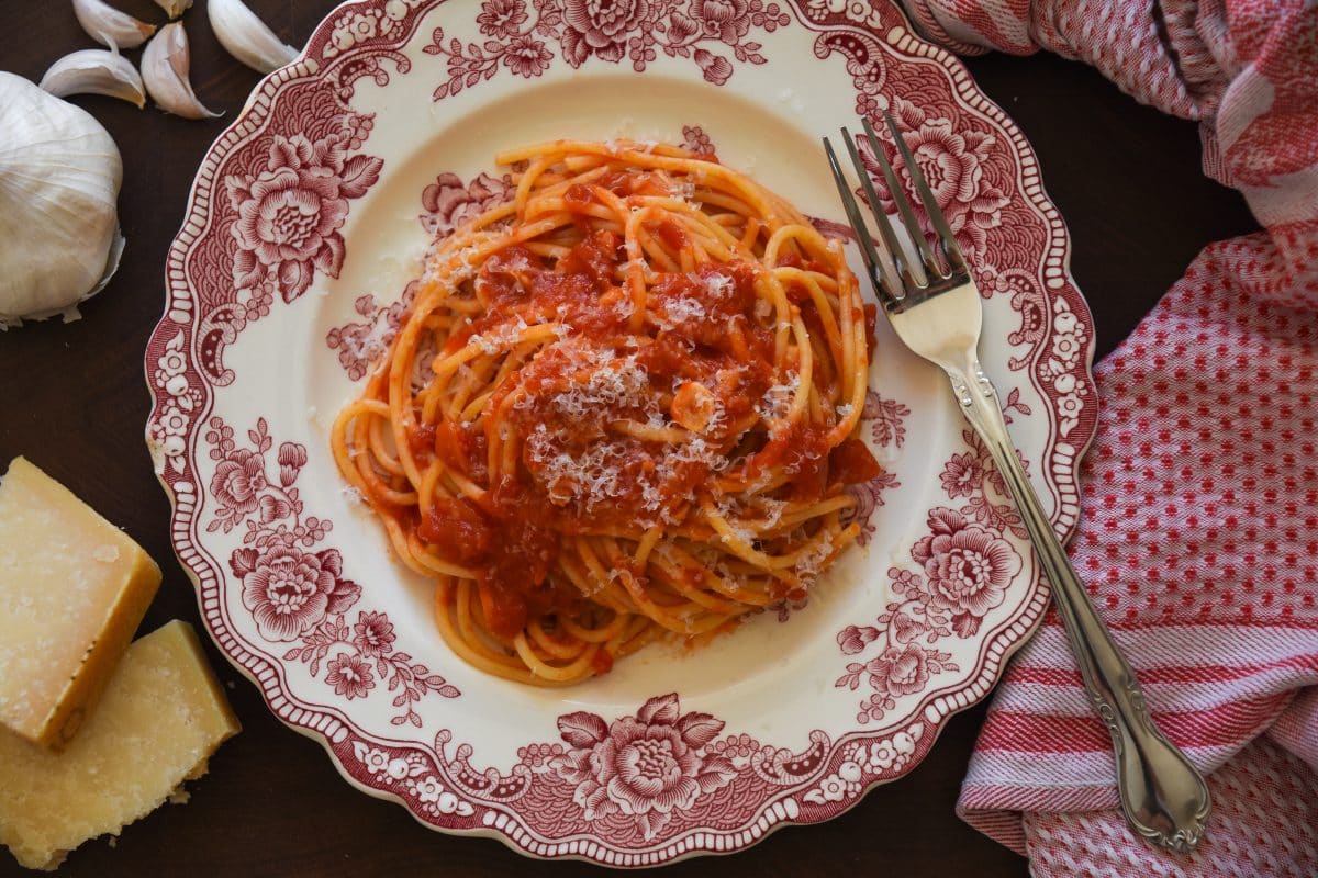 A plate of spaghetti marinara with a fork surrounded by a kitchen towel, garlic, and parmesan cheese. 